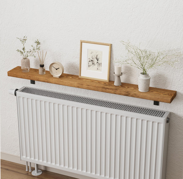 a wooden shelf mounted on a wall, with a white radiator below it. On the shelf, there are several decorative items, including a framed picture, a clock, a vase with flowers, and a small plant.