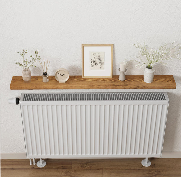 a white radiator mounted on a wall, with a wooden shelf above it holding various decorative items such as a clock, a picture frame, and two potted plants.