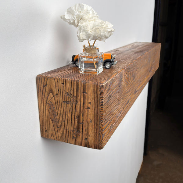a wooden shelf mounted on a wall, with a vase containing white flowers placed on top of it. The shelf has a rustic, natural finish and is made of wood. There is also a small toy car placed on the shelf.
